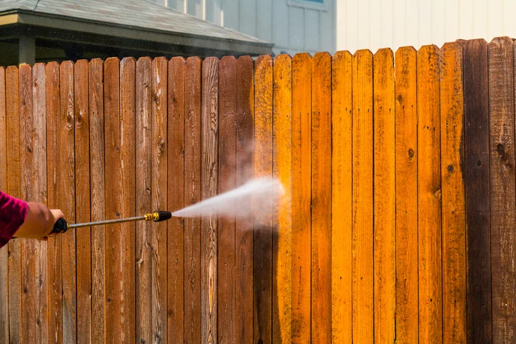Pressure washing wooden fence with high-pressure spray, showing cleaned versus weathered wood sections